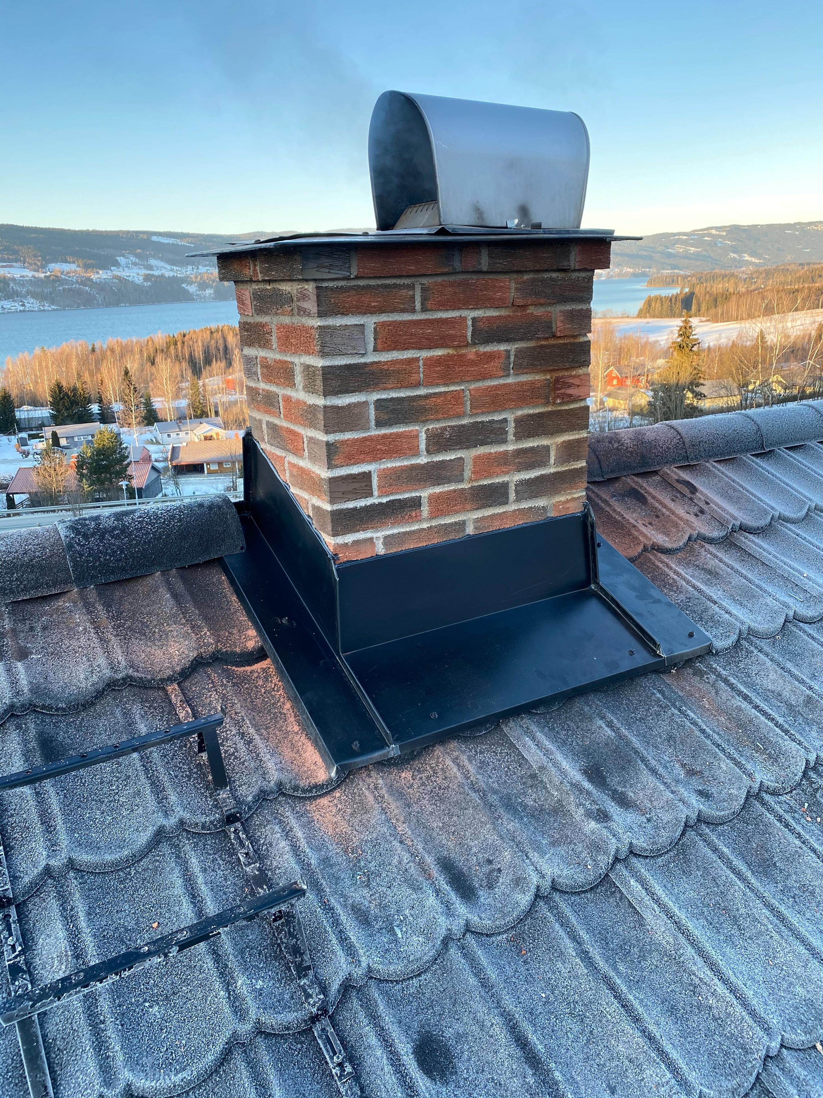 Mursteinpipe på taket med utsikt over innsjø og landskap i bakgrunnen.