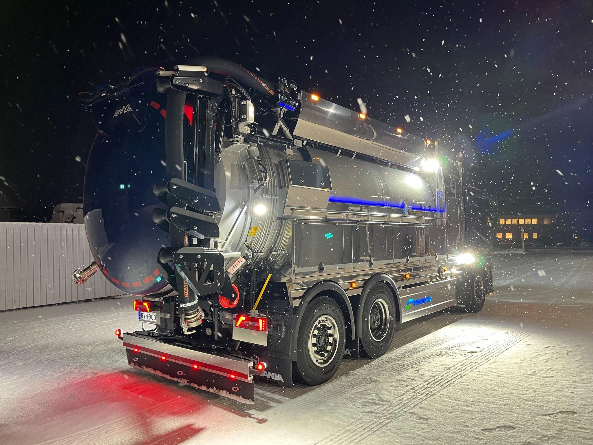 En stor søppelbiler med lys som lyser opp snøen om natten, står parkert i et vinterlandskap.