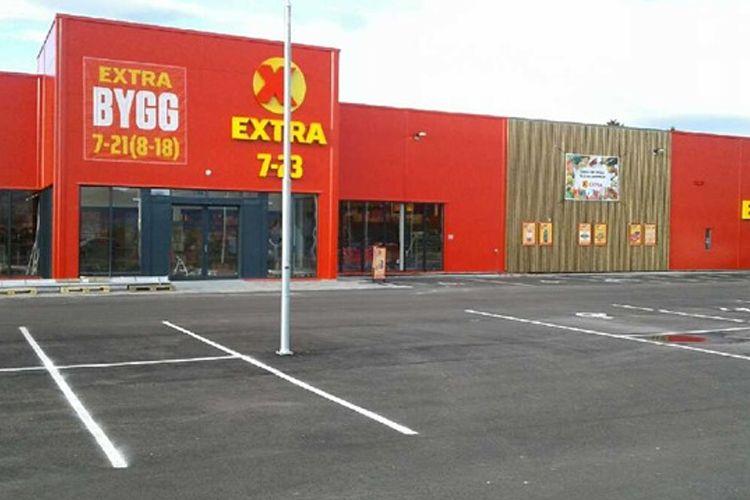 En stor byggevarebutikk med rød fasade og parkeringsplass foran. Skilt viser åpningstider.