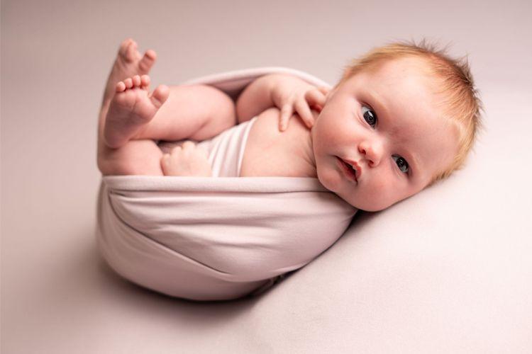 Nyfødt baby med rødt hår, pakket inn i en myk, lys stoff, ligger på en hvit bakgrunn.