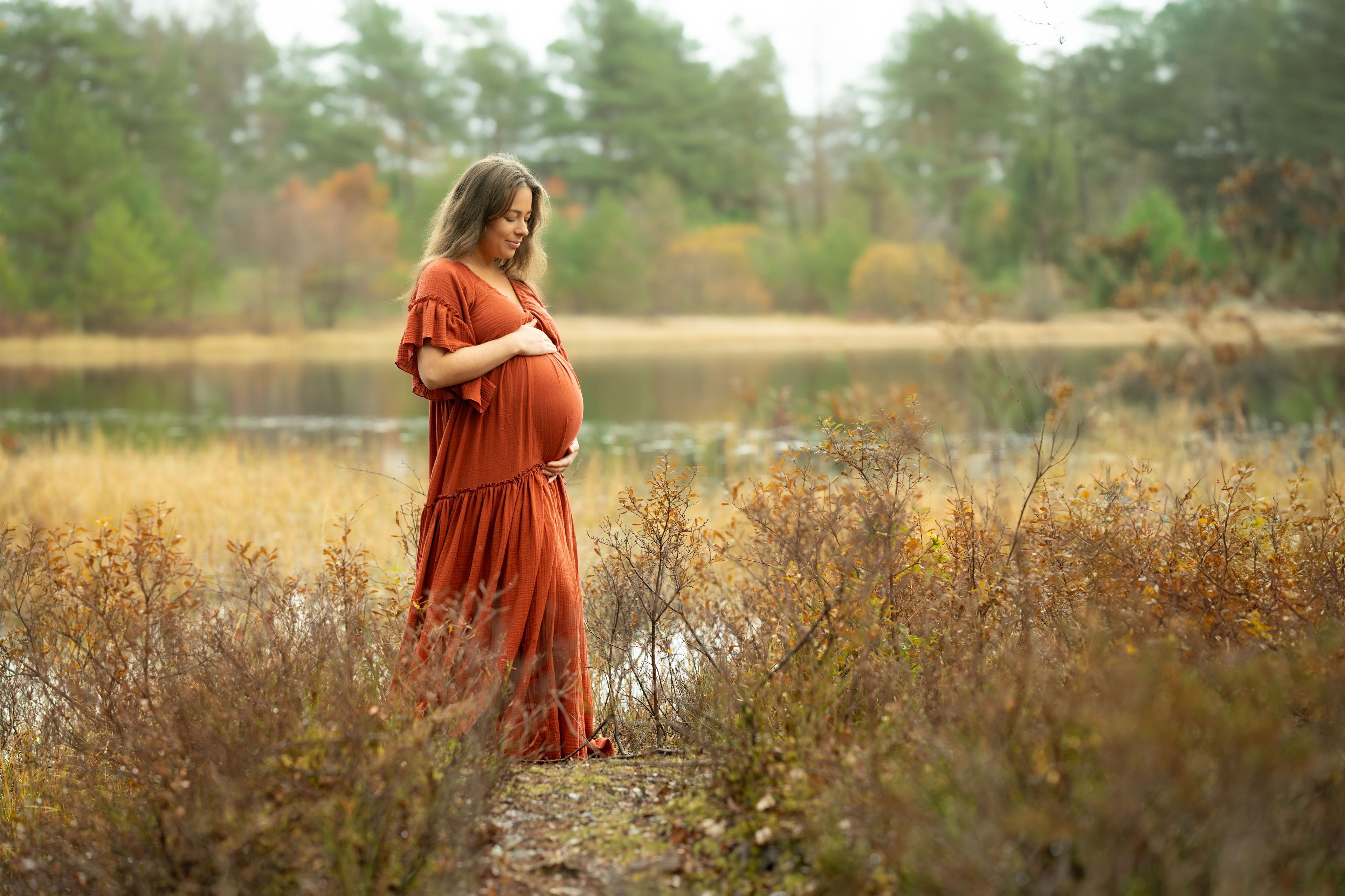 Gravid kvinne i rød kjole står ved vannkanten med naturen i bakgrunnen.