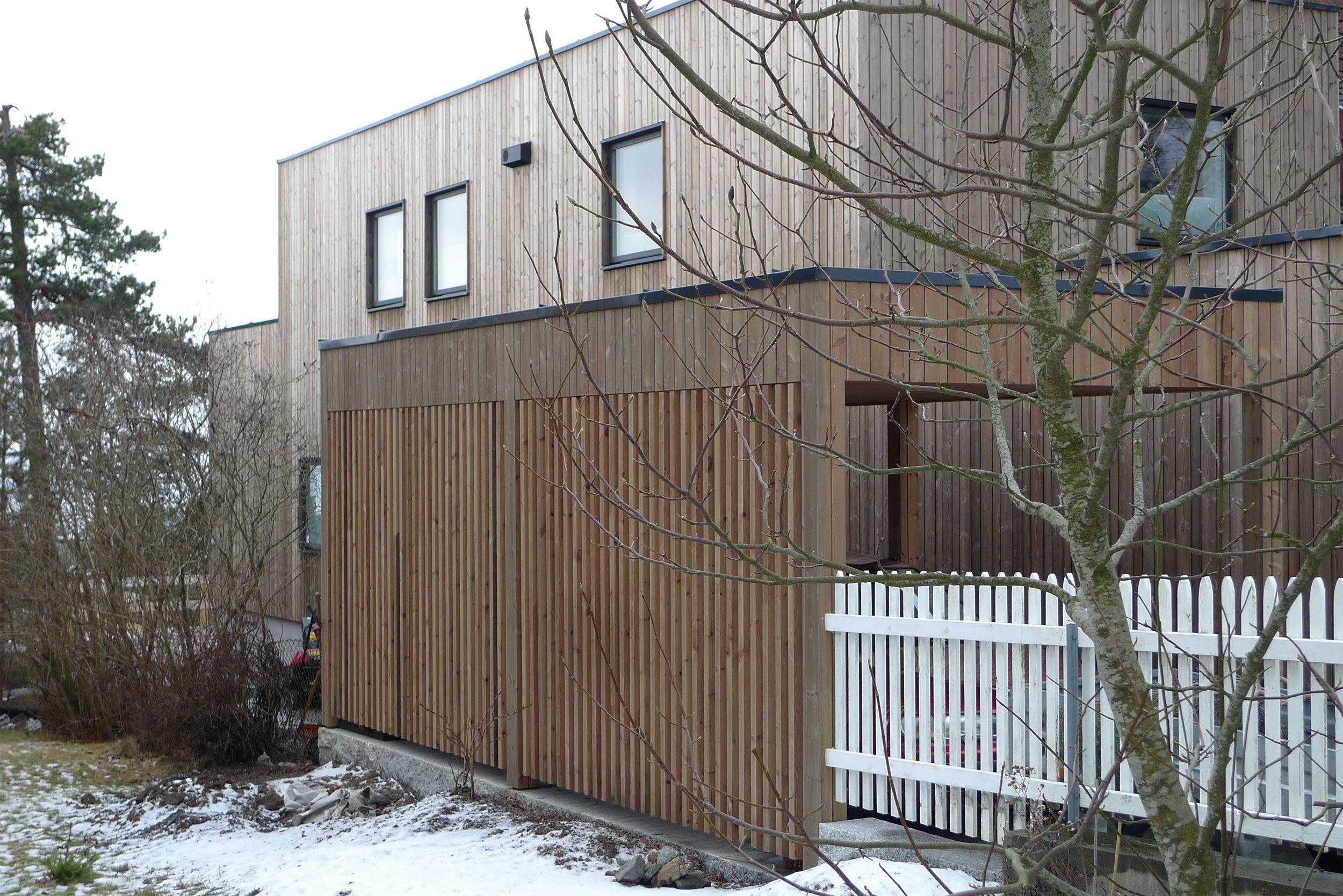 Moderne hus med trepanel og hvit gjerde, omringet av snø og trær.