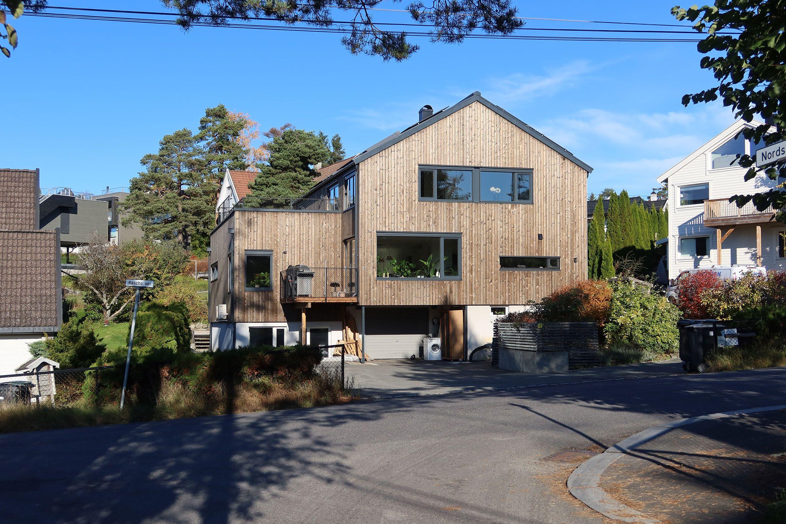 Moderne trehus med store vinduer, beliggende i et boligområde med trær og grønt rundt.