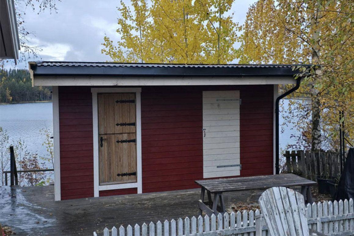 Rødt hytte med tre dører ved vannet, omgitt av høstfargede trær og en hvit plankehekk.