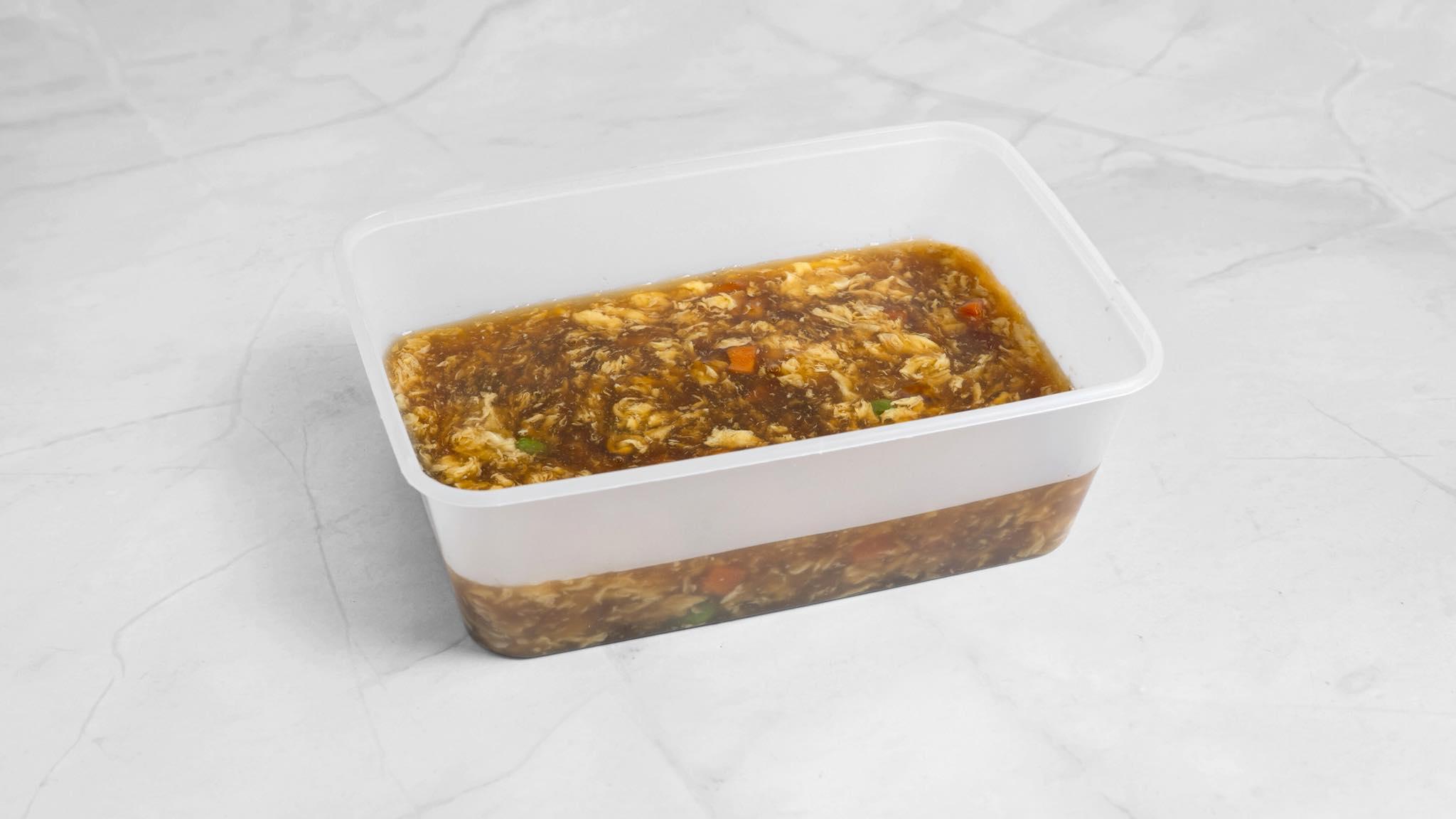 En plastbeholder med brun saus som inneholder grønnsaker og krydder på en lys marmoroverflate.