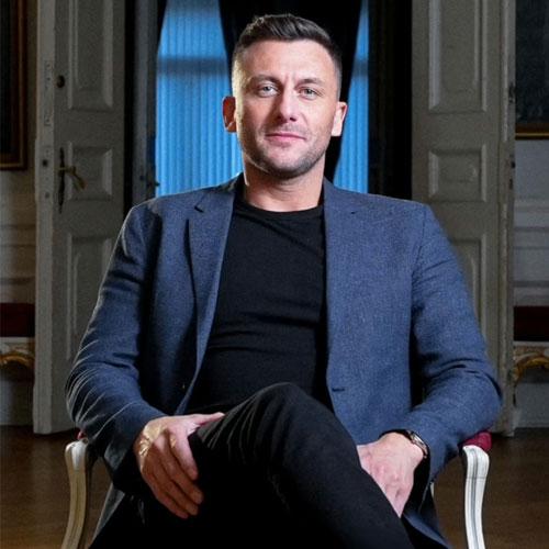 En mann sitter i en stol med armene krysset, iført en blå blazer og svart genser, i et rom med vinduer.