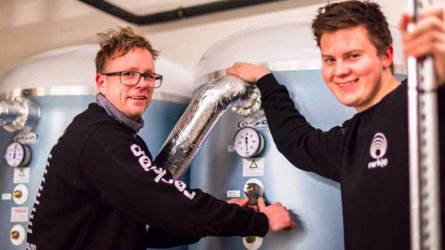 To personer som arbeider med utstyr i et laboratorium, med store tanker og måleinstrumenter i bakgrunnen.