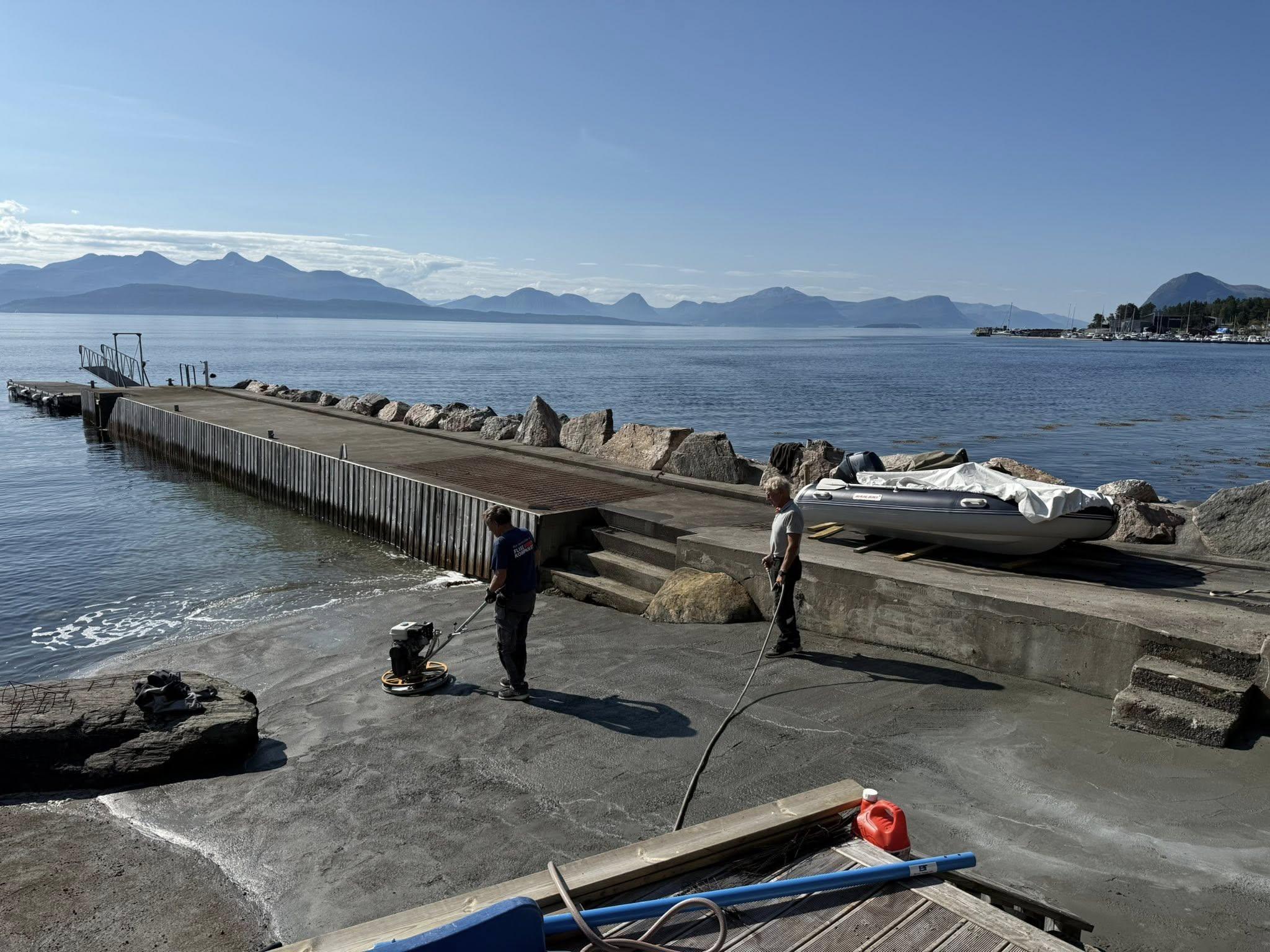 To menn står på en brygge ved sjøen, med fjell i bakgrunnen og en båt på land. Solfylt vær.