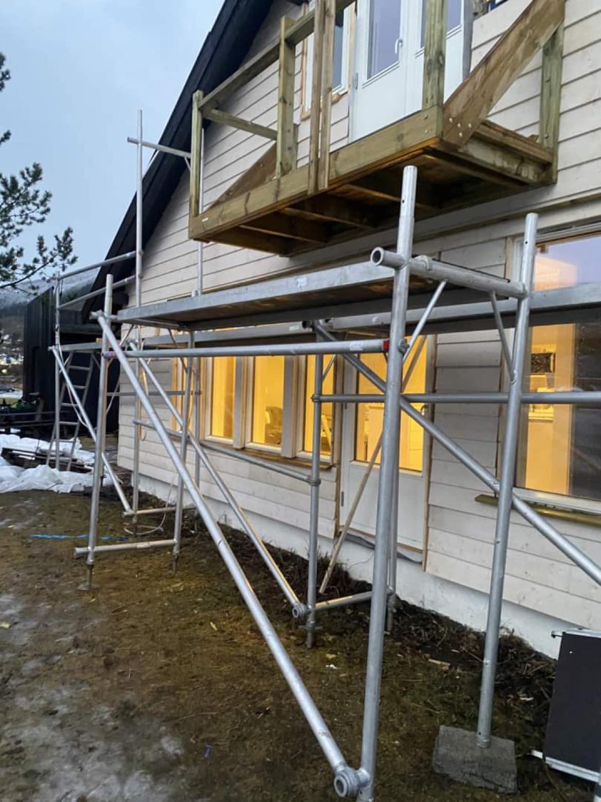Stillase ved en bygning med vinduer og en balkong, klar for renovering. Snø og grus på bakken.