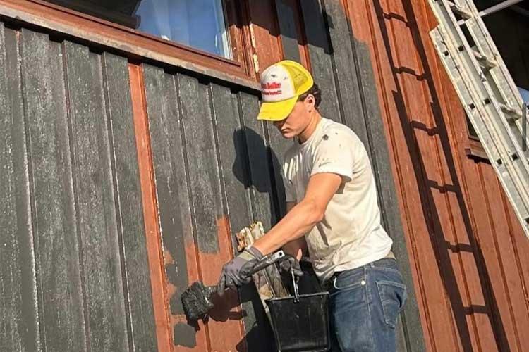 En ung mann maler en vegg med svart maling, iført hansker og en caps, med en stige i bakgrunnen.