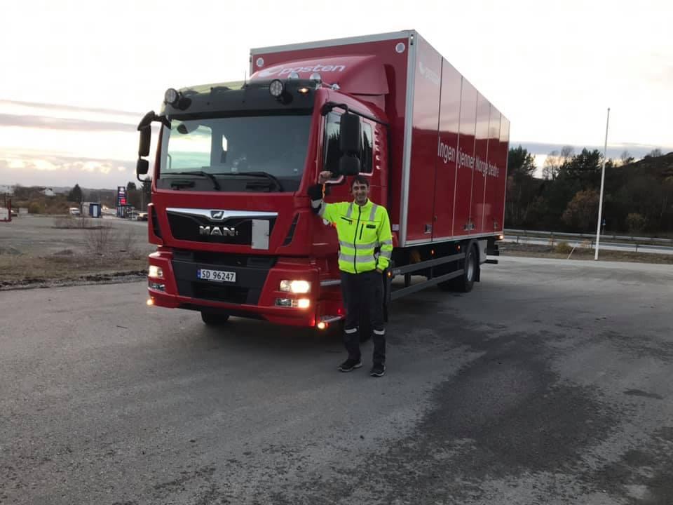 En sjåfør står ved siden av en rød MAN lastebil på en parkeringsplass i solnedgang.