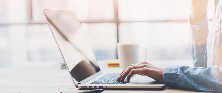 En person skriver på en laptop med en kopp kaffe ved siden av, i et lyst rom med vinduer.