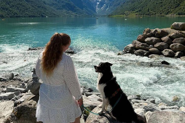 Kvinne med langt hår står ved elvebredden med en hund, med fjell og vann i bakgrunnen.