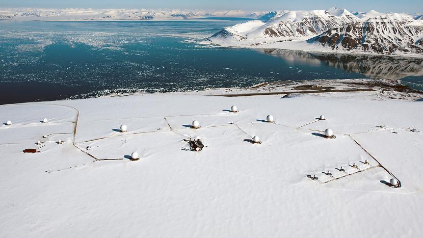 Oversiktsbilde av en snødekt forskningsstasjon med satellittantenner i Arktis, omgitt av fjell og hav.