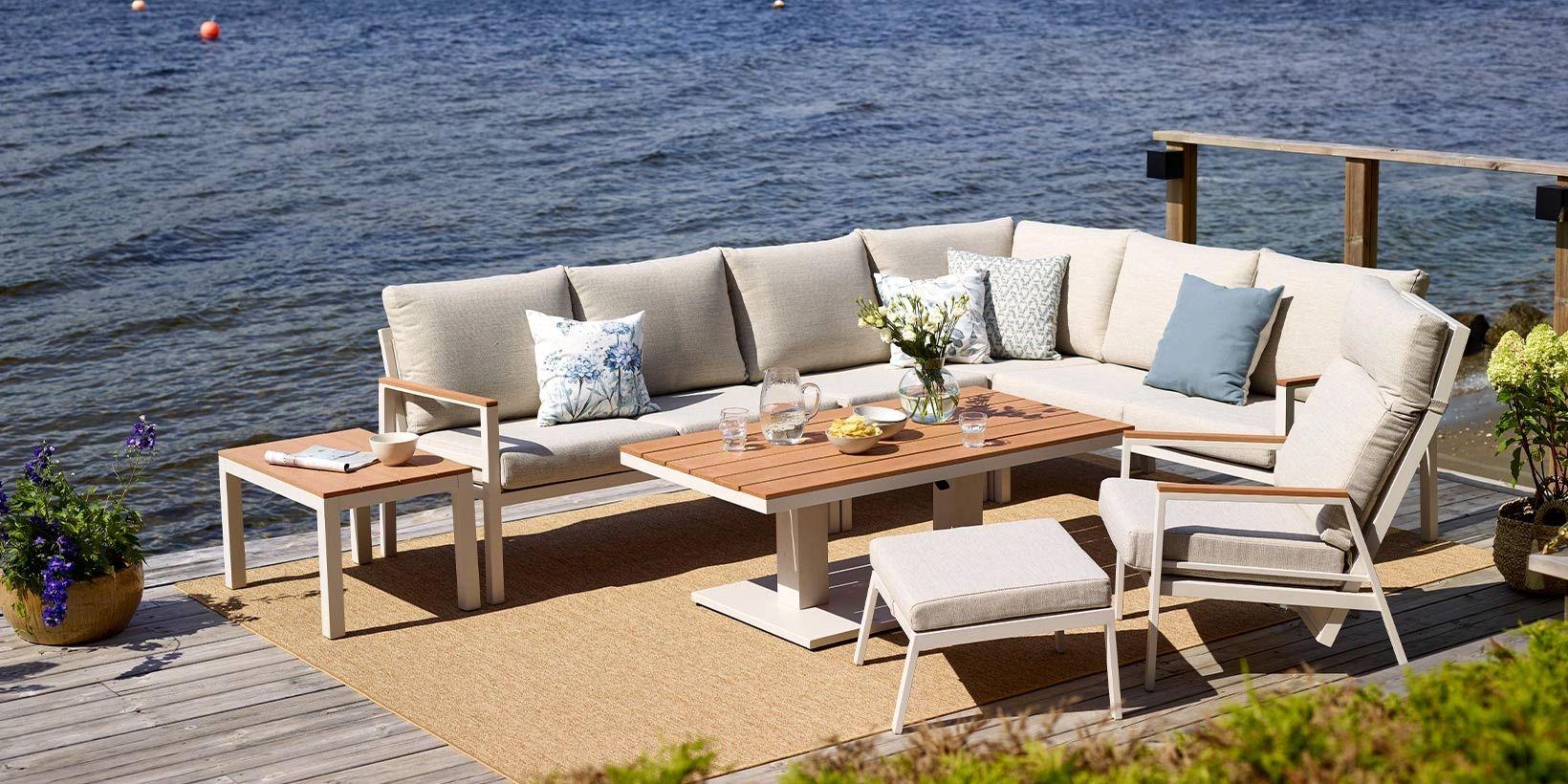 Uteplass med en stor sofa, bord og stoler ved vannet, dekorert med puter og blomster.