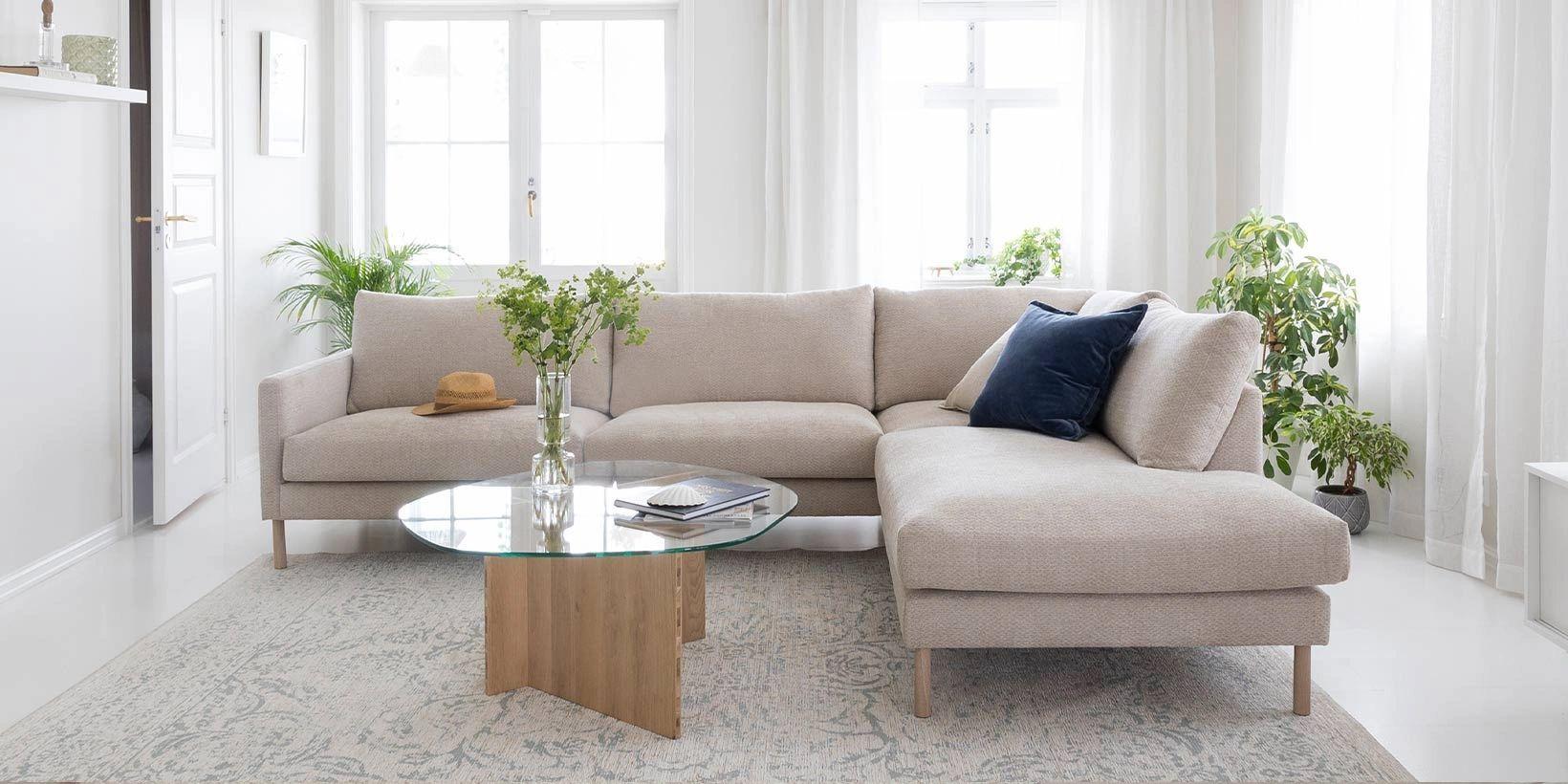Lys stue med en beige sofa, et glassbord og grønne planter i vinduskarmen.