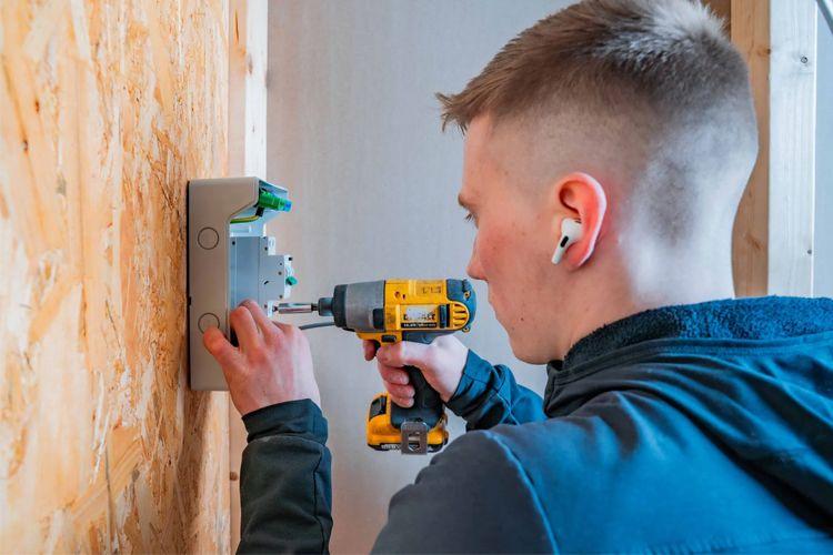 En ung mann bruker en drill for å installere en enhet på en vegg av trepanel.