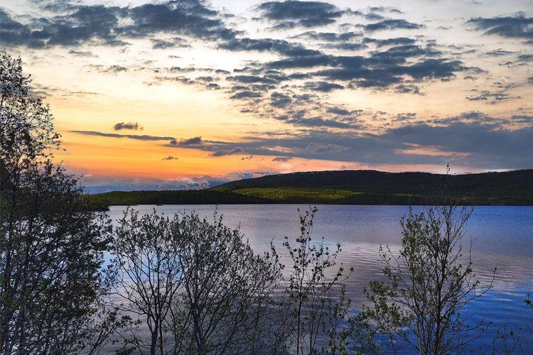 Solnedgang over en innsjø med trær i forgrunnen og fjell i bakgrunnen. Himmel med skyer i varme farger.