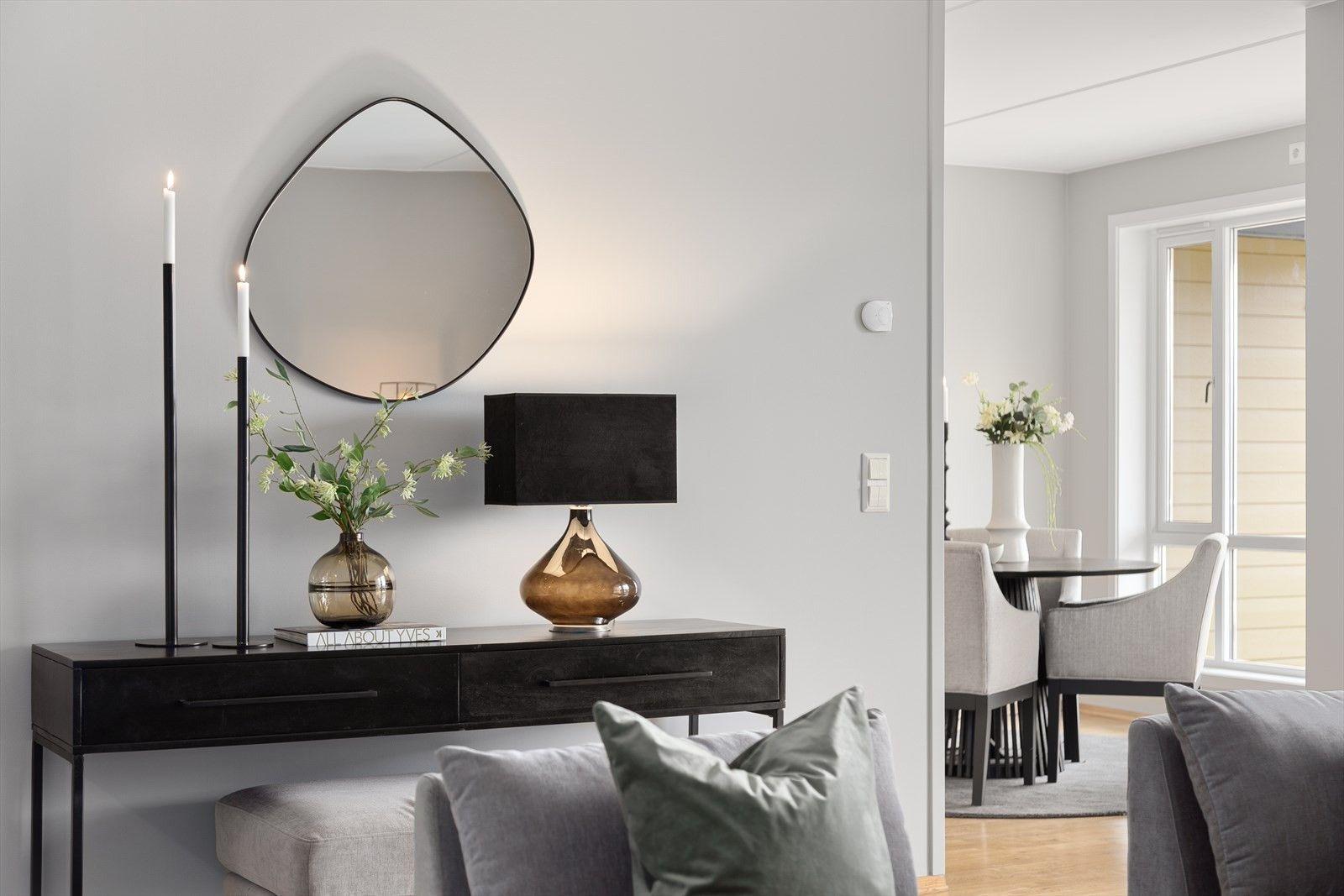 Moderne stue med svart konsollbord, speil, lysestaker og en elegant lampe, samt en spiseplass i bakgrunnen.