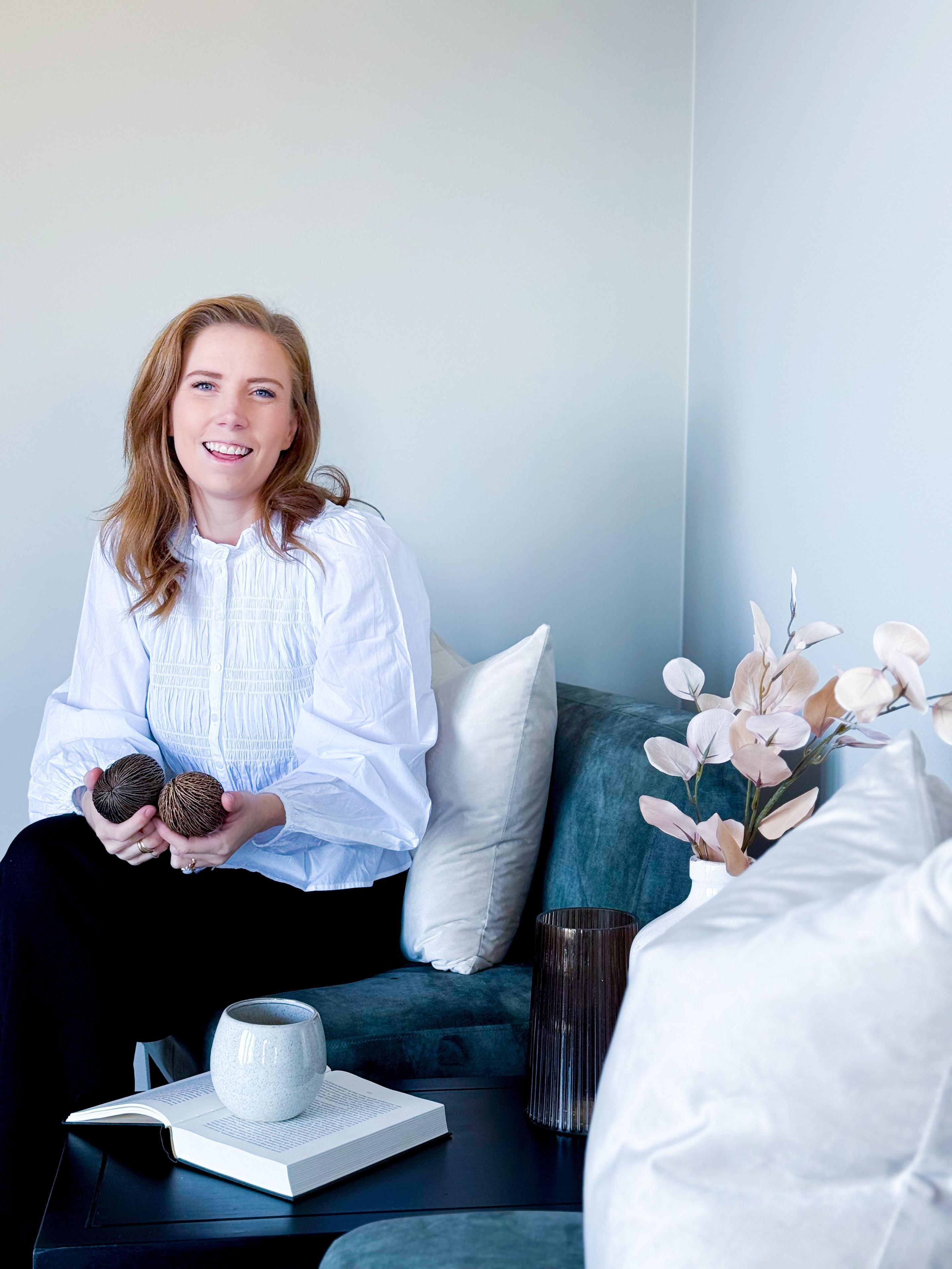 Kvinne med langt hår sitter på en sofa, holder to dekorative gjenstander, omgitt av puter og en vase med tørkede blomster.