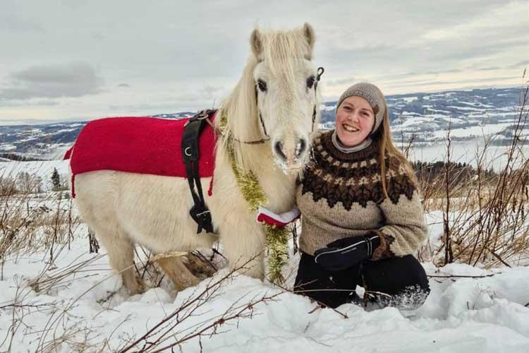Kvinne med et hvitt pony i snøen, iført rød dekken, med vakker vinterlandskap i bakgrunnen.