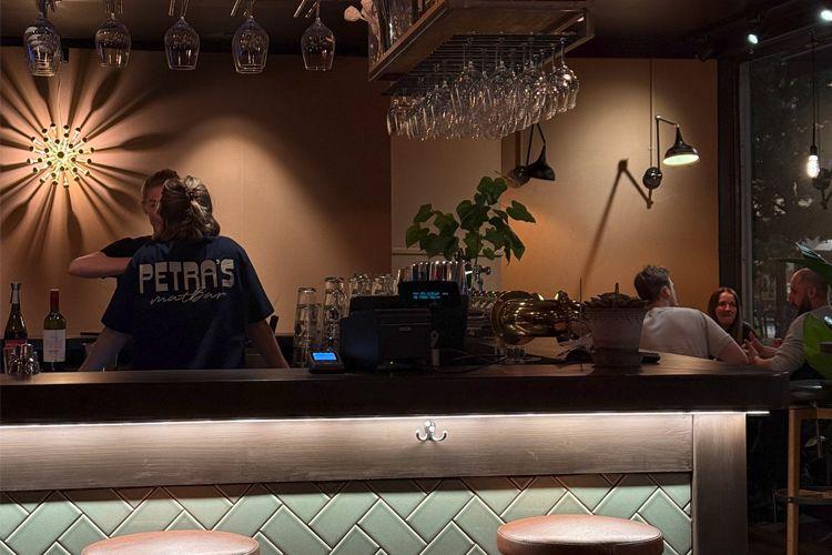 En koselig bar med bartendere som serverer gjester, glass og flasker på hyllen, og folk som sitter ved bordene.
