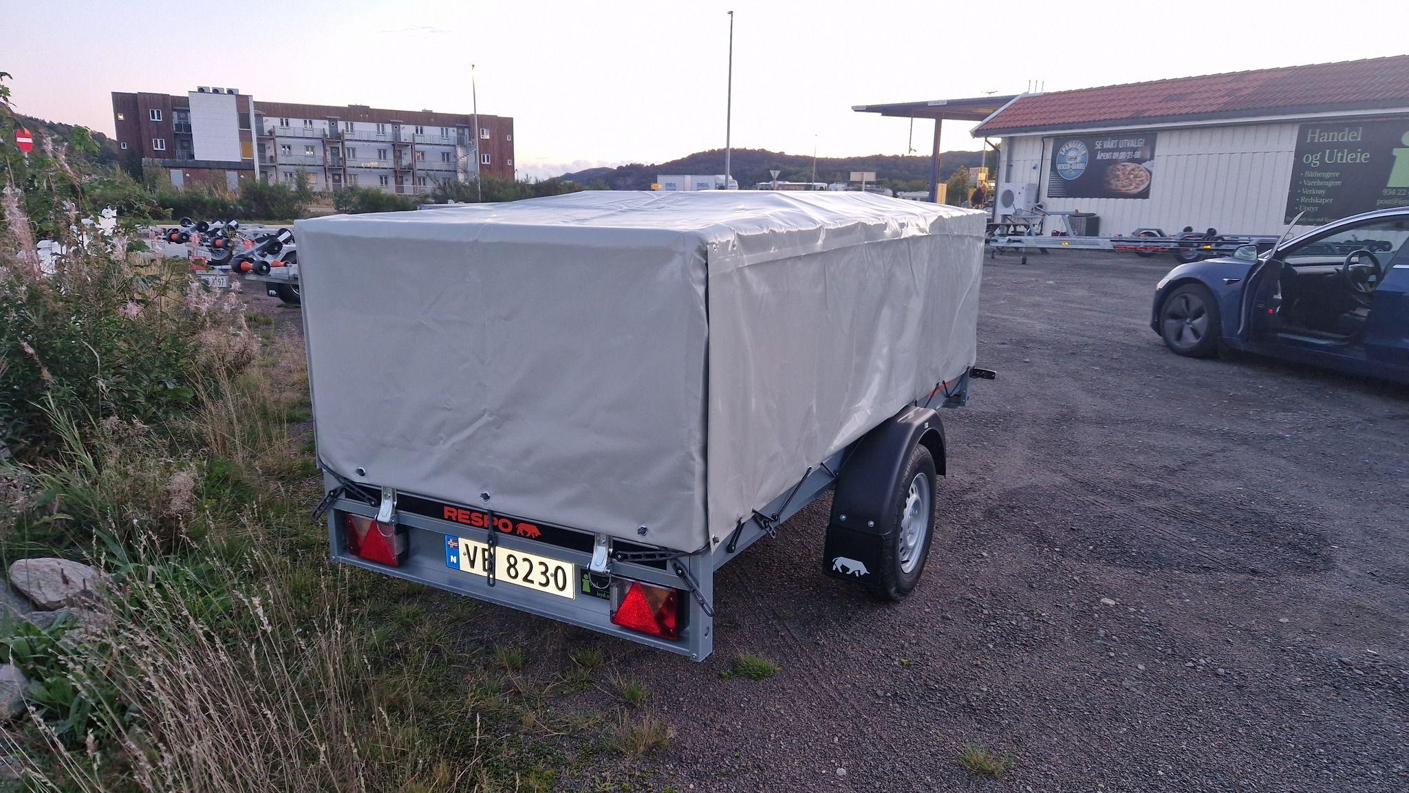 En tilhenger med grå presenning parkert på en grusplass, med bygninger i bakgrunnen.