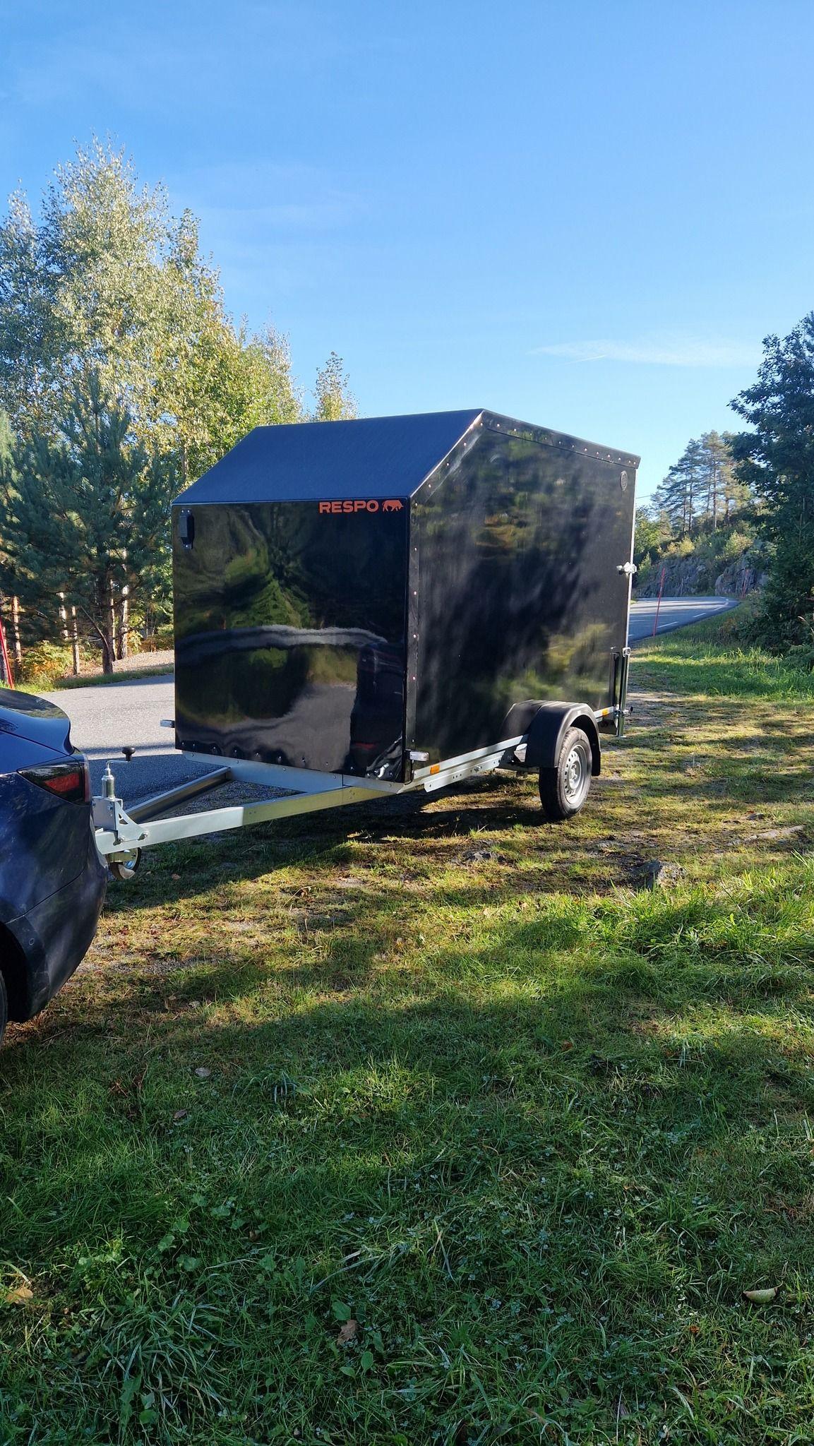 En svart tilhenger parkert på gresset ved veien, med trær i bakgrunnen og klart blå himmel.