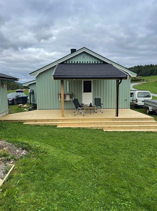 En moderne hytte med terrasse, stoler og bord, omgitt av grønt gress og landskap under en skyet himmel.