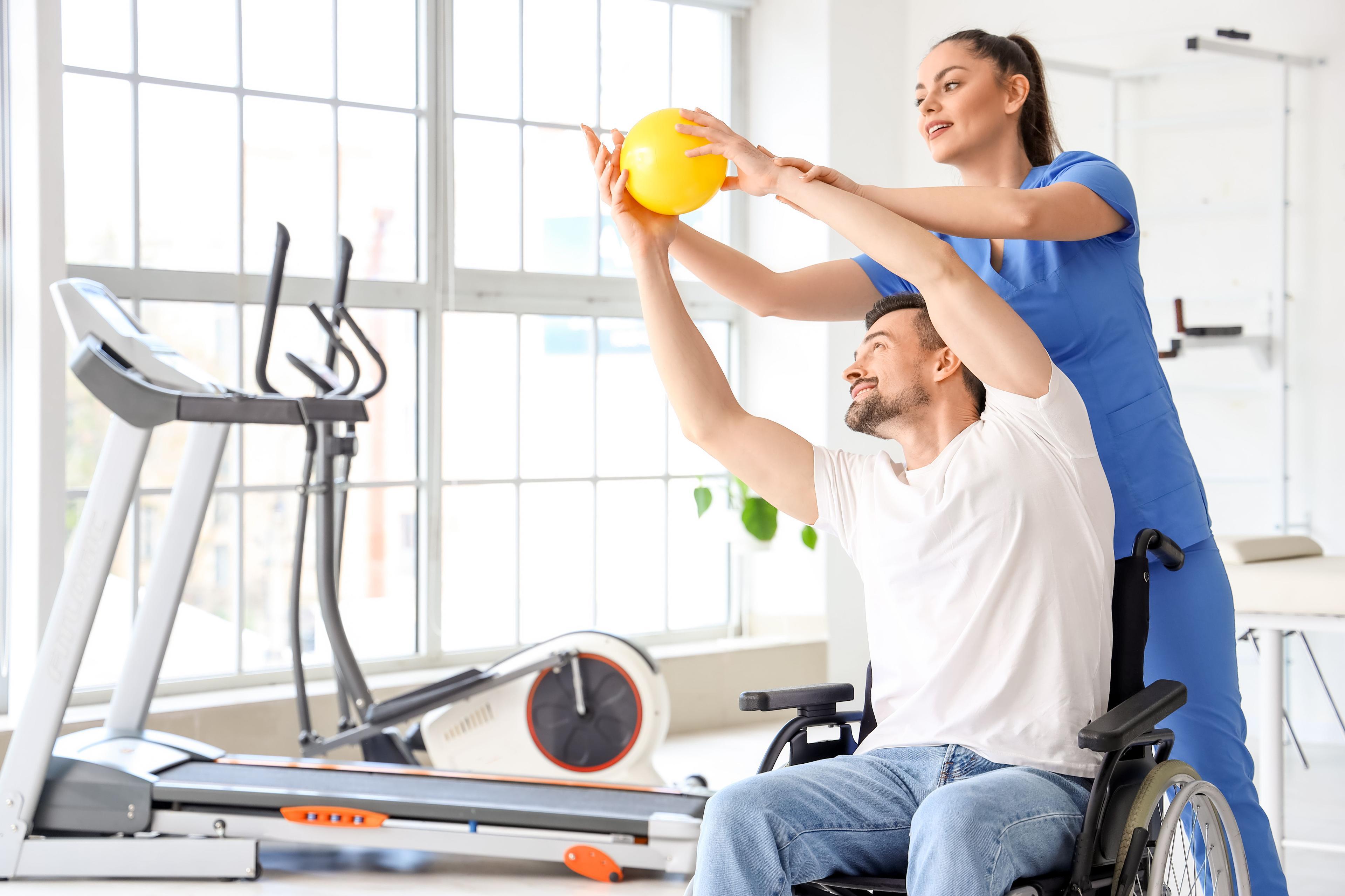 En kvinne hjelper en mann i rullestol med trening ved hjelp av en ball i et moderne treningsrom.