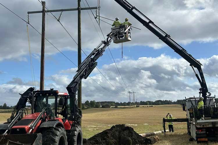 To personer arbeider med strømledninger fra en heisekran, mens en traktor står i feltet.