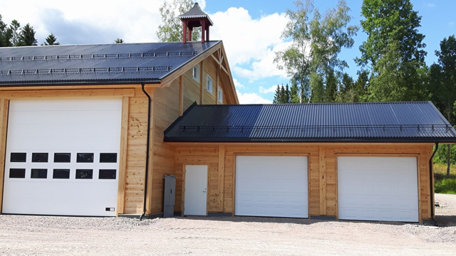 Moderne trehus med to garasjeporter og svart tak, omgitt av trær og blå himmel.