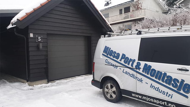 En varebil fra Mjøsa Port & Montasje parkert foran en svart garasje i snødekt landskap.