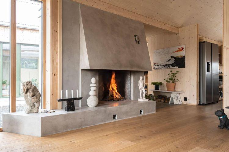 Moderne stue med peis i betong, brennende ild, kunstverk og store vinduer som slipper inn naturlig lys.