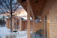 Moderne hytte med trepanel, snødekt landskap og solnedgang i bakgrunnen. Stoler på verandaen.
