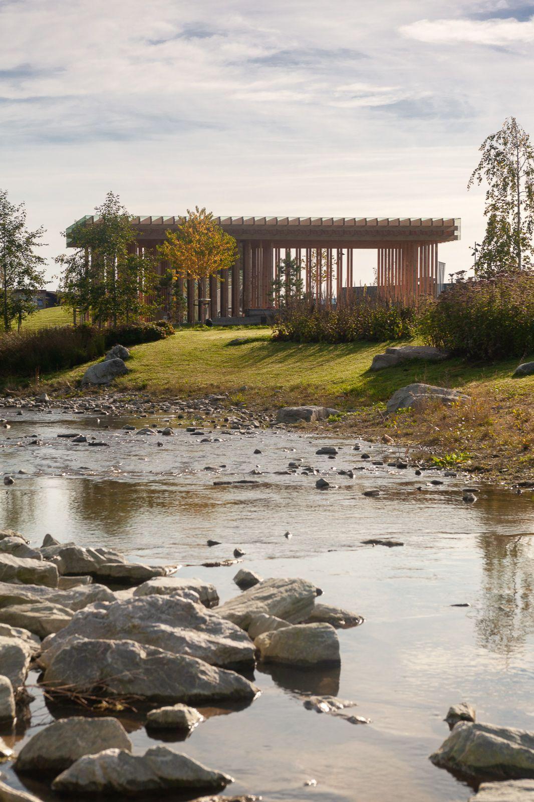 En elv med steiner i forgrunnen og en moderne bygning i bakgrunnen, omgitt av trær og grønt gress.