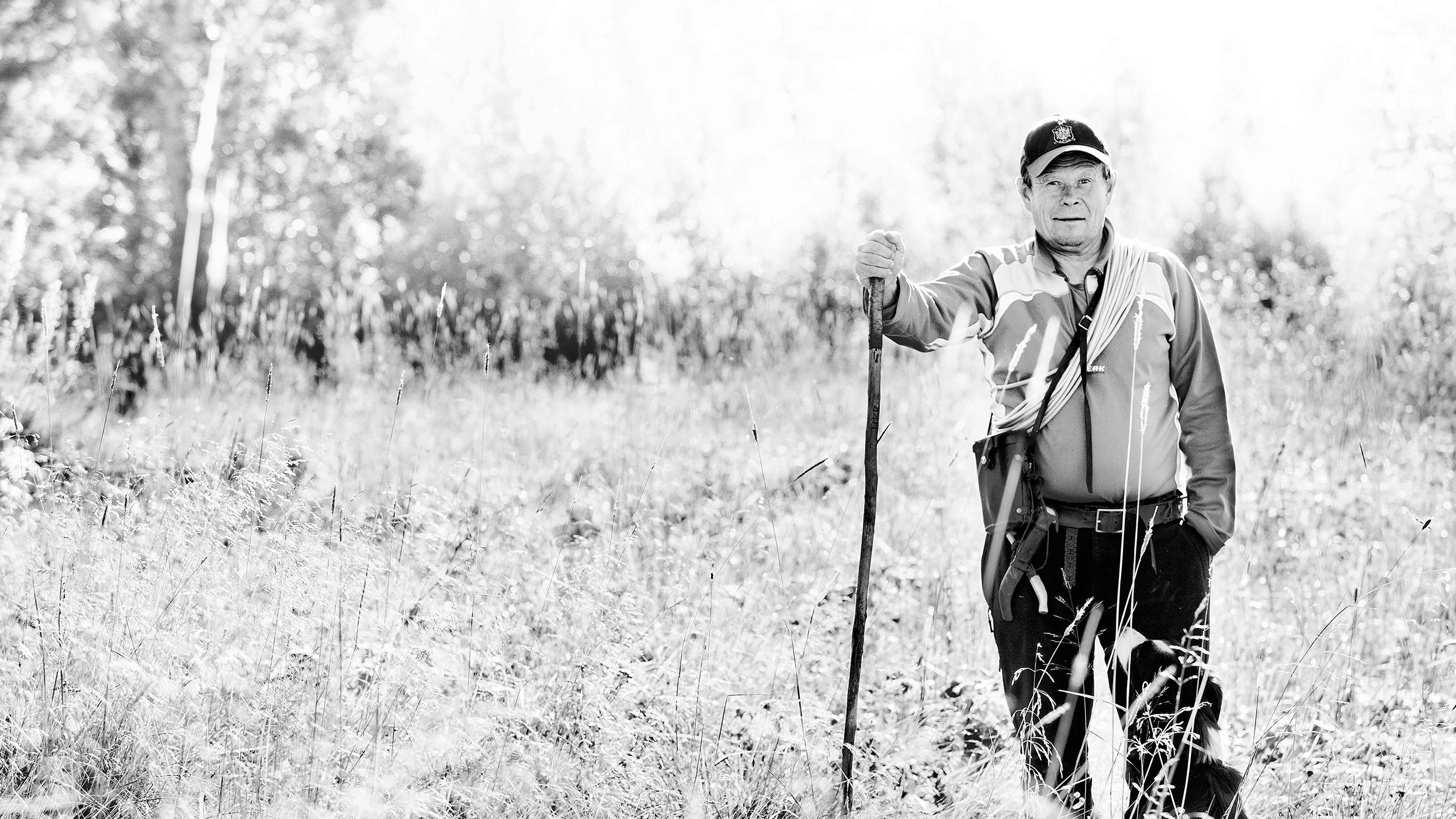 En eldre mann står i en eng med en stav, iført jakke og caps, omgitt av trær og høyt gress.
