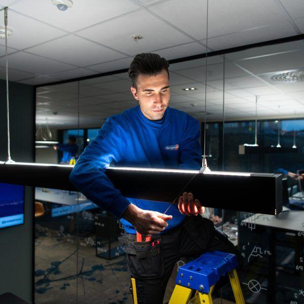 En elektriker som monterer en lysarmatur på en stige i et moderne kontorlandskap.