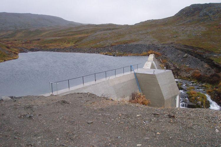 En demning med vann og fjellbakgrunn, omkranset av høstfarger og grusete terreng.