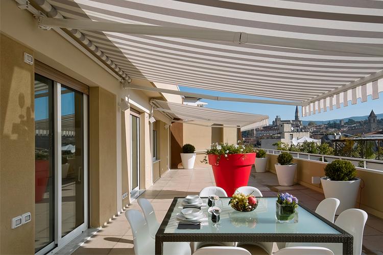 Moderne terrasse med glassbord, hvite stoler og blomsterpotter, med utsikt over byen og blå himmel.