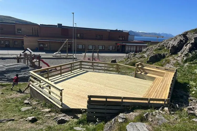 Uteområde med tredekk og rekkverk, nær en skolebygning og lekeplass i solfylt vær.