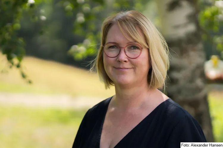 En kvinne med brunt hår og briller står ute i naturen, med trær og grønt landskap i bakgrunnen.