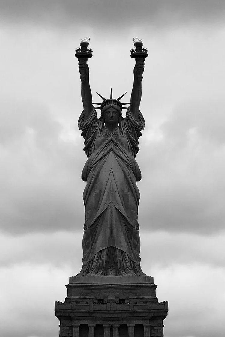 Statue of Liberty i svart-hvitt, med armene hevet og flammer i hendene, mot en skyet himmel.