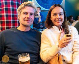 To personer sitter sammen i en bar, smilende og holder glass med drikke. Bakgrunnen viser en livlig atmosfære.