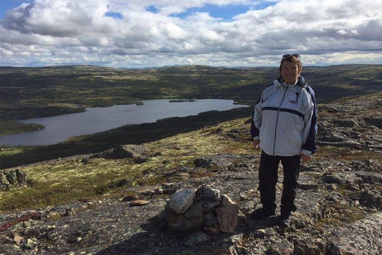 En person står på en fjelltopp med utsikt over en innsjø og fjellandskap under en skyet himmel.