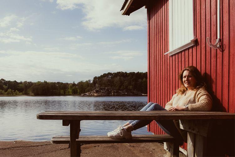 Kvinne sitter på benk foran rød hytte ved vannet, med solfylt himmel og grønn natur i bakgrunnen.