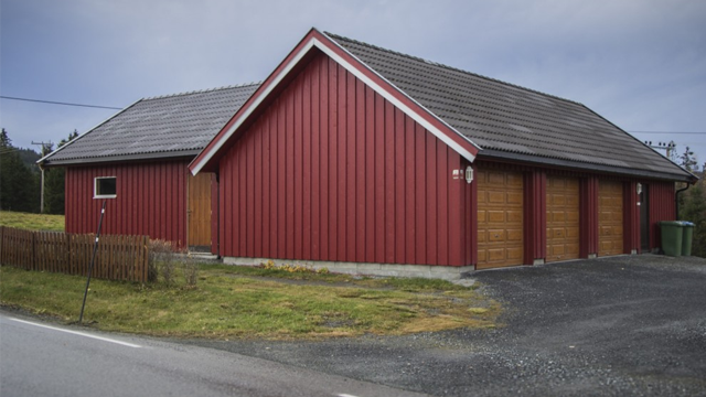 Rødt hus med garasje og tre dører, beliggende ved veien i et landlig område med grønt gress.