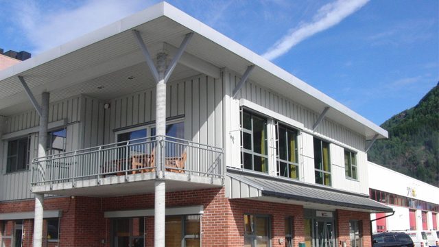 Moderne bygning med balkong, store vinduer og grå fasade, omgitt av fjell og blå himmel.