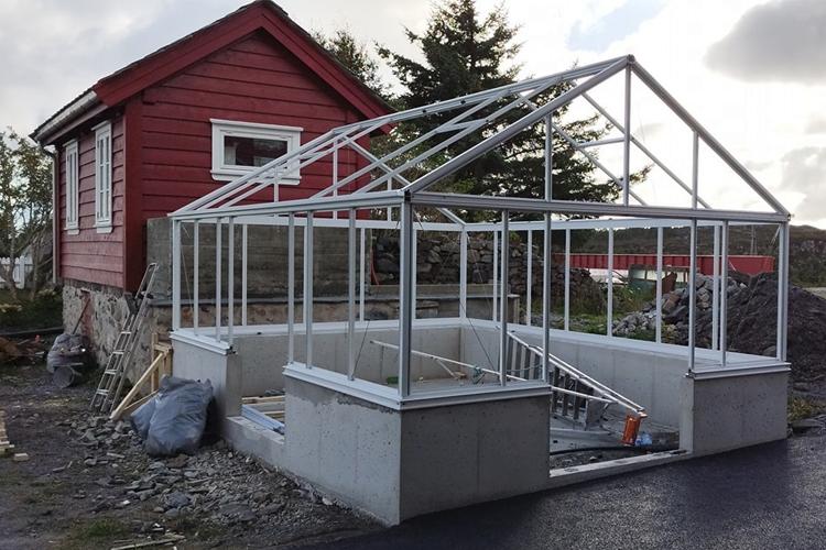 Rødt hus med et påbygg av glassveranda i konstruksjon, omgitt av stein og trær.