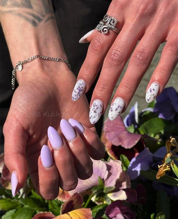To hender med lange negler, en hånd med lys lilla neglelakk og en annen med hvit neglelakk med blomsterdesign.